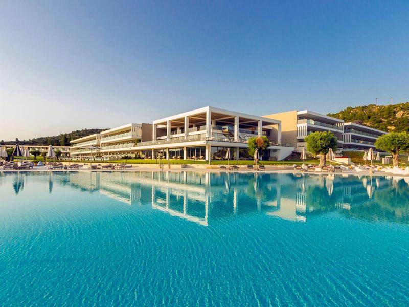 Moderner Hotelkomplex mit großem Pool, klar blauem Wasser und grünem Hügel im Hintergrund.