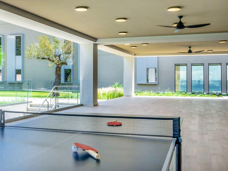 Tischtennisplatte unter einem überdachten Bereich mit Ventilatoren, im Hintergrund Grünfläche und Gebäude.