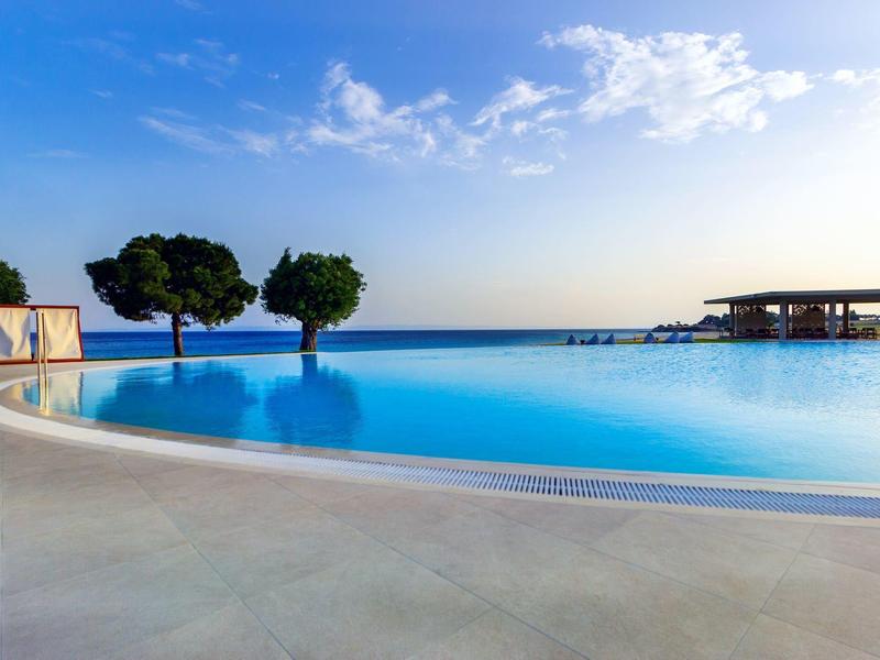 Großer, blauer Infinity-Pool mit klarer Wasseroberfläche, Bäumen und bewölktem Himmel im Hintergrund.