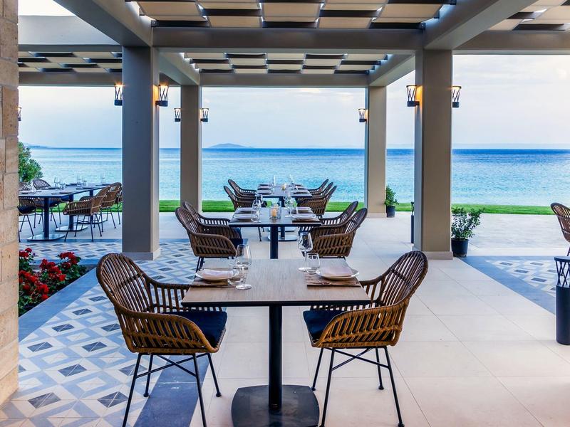 Terrassenrestaurant mit Tischen, Stühlen und Blick auf das blaue Meer unter klarem Himmel.