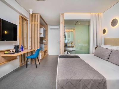 Modern hotel room with double bed, desk, blue chair, and glass bathroom.