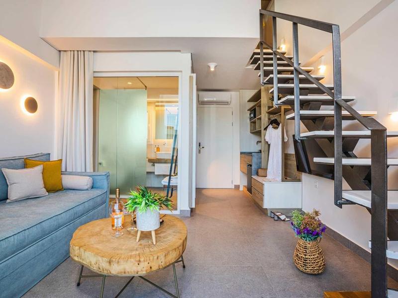 Chambre d'hôtel moderne avec escalier, canapé, cloison en verre et coin salon aux tons neutres.