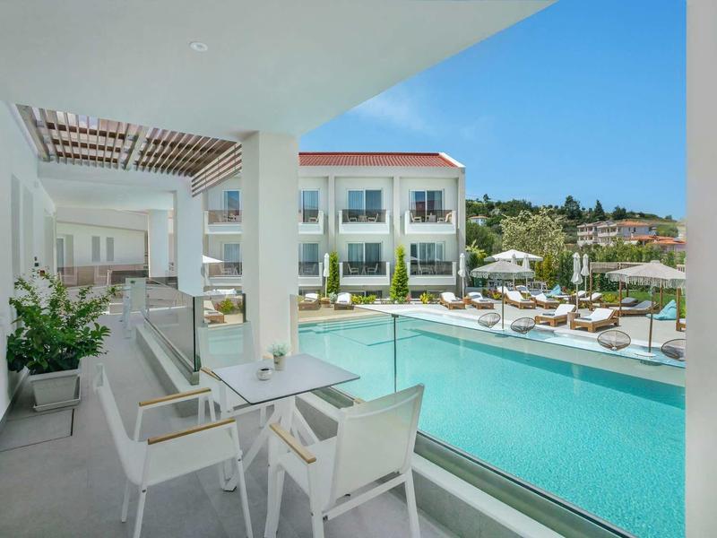 Vue sur une piscine extérieure moderne avec des chaises et tables blanches sur une terrasse et des bâtiments d'hôtel en arrière-plan.