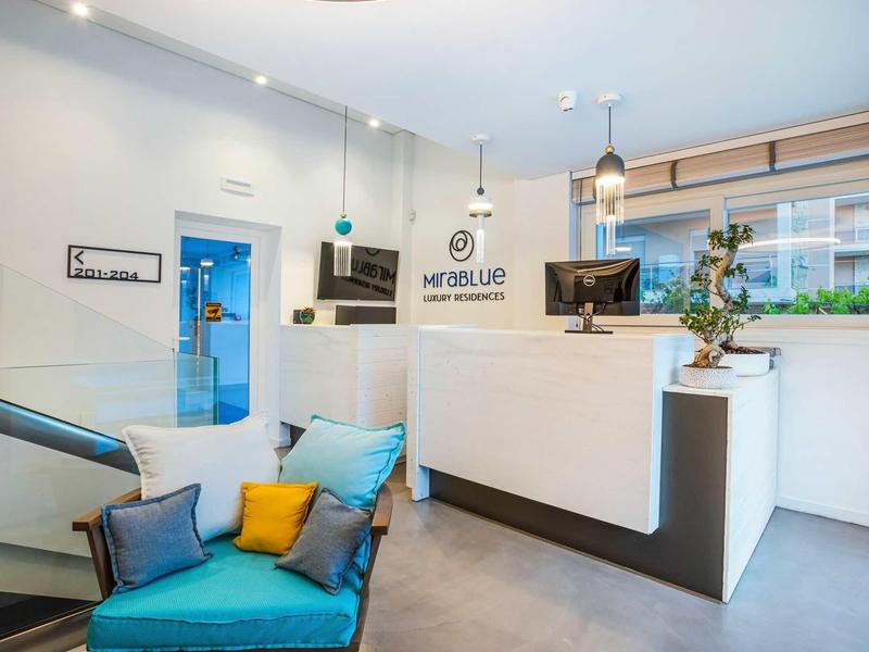 Hall d'hôtel lumineux avec réception moderne, canapé aux coussins colorés et grande fenêtre.