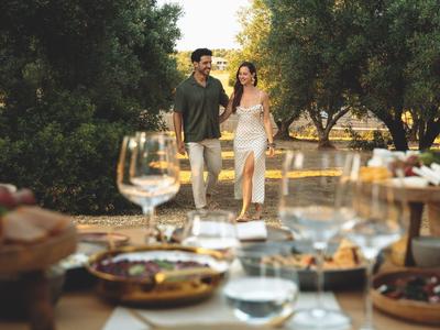Paar spaziert auf einem mit Bäumen gesäumten Weg, davor ein gedeckter Tisch mit Essen und Wein.