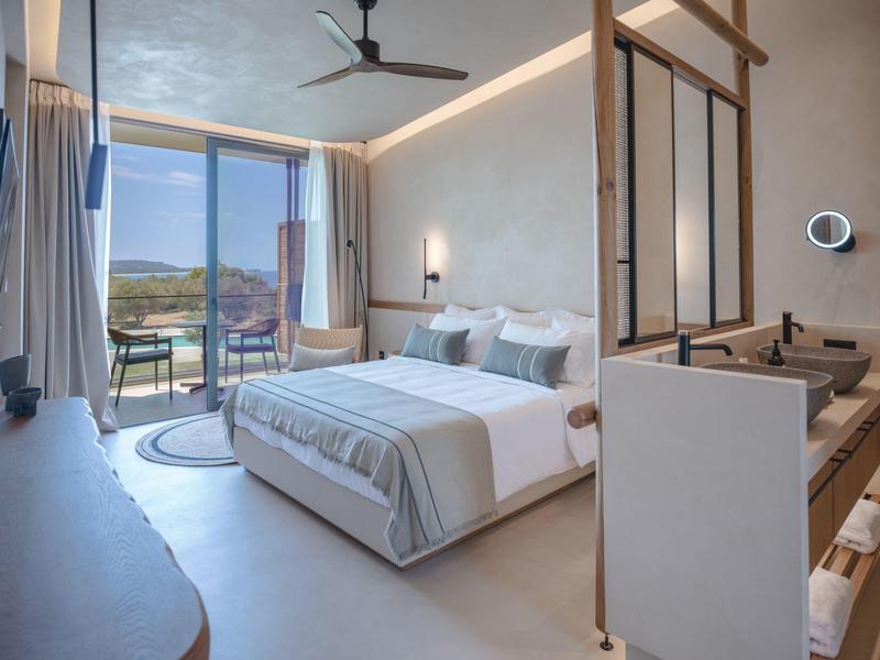 Modern hotelkamer met groot bed, balkon met uitzicht op heuvels en open badkamergedeelte.