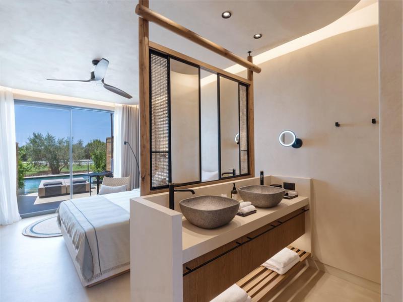 Modern hotelkamer met tweepersoonsbed, grote ramen en dubbele wastafels in de badkamer.