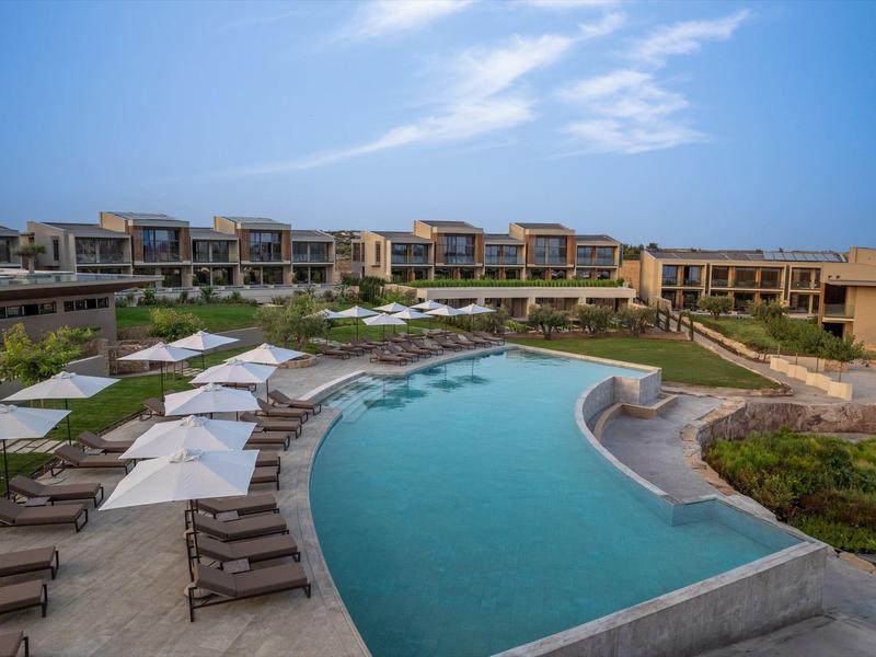 Modern hotelcomplex met groot gebogen buitenzwembad, ligstoelen en parasols onder een blauwe lucht.
