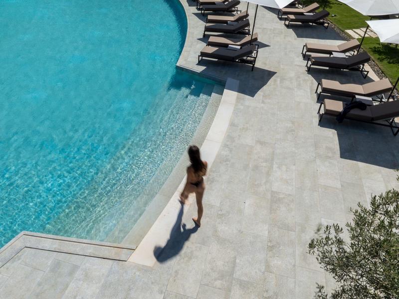 Vrouw stapt in het water bij het hotelzwembad op een lichtgrijze stenen vloer, omgeven door ligbedden op gras.