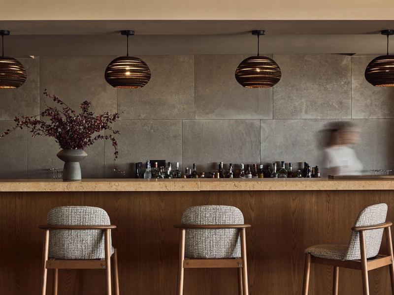 Drie stoelen bij een houten bar met moderne hanglampen en een wazige ober op de achtergrond.