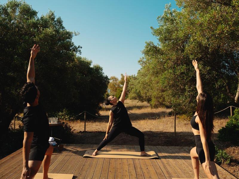 Drie personen doen yoga op een houten platform buiten bij zonnig weer.