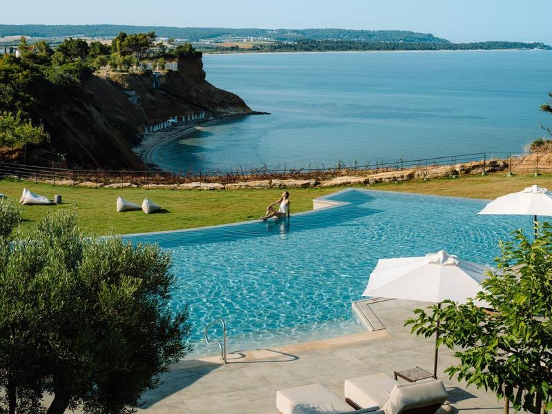 Infinity zwembad met ligstoelen met uitzicht op rustige baai en kliffen onder heldere lucht