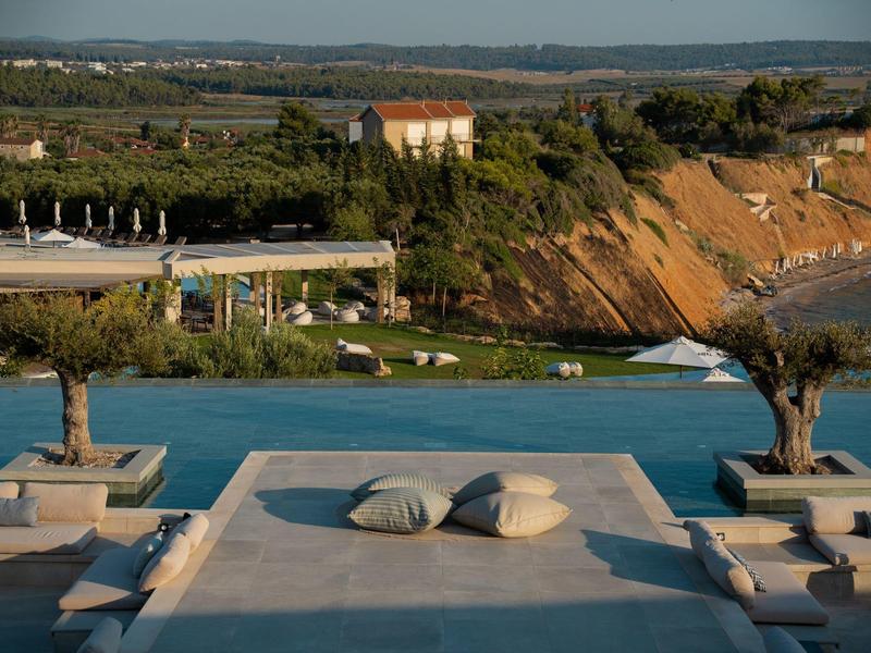 Blick von einer Sonnenliege auf einen Pool mit Palmen und Hügeln im Hintergrund.