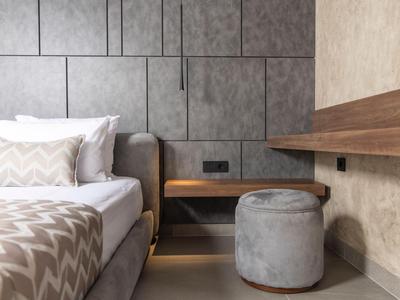 Modernes Hotelzimmer mit grauem Bett, Nachttisch und rundem Hocker vor strukturierter Wand.