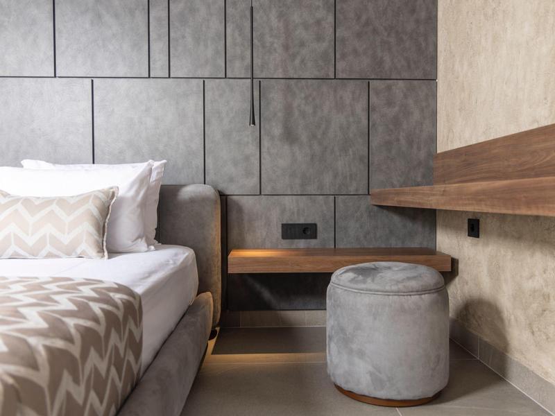 Nowoczesna sypialnia z szarym łóżkiem, tapicerowanym stołkiem i drewnianym stolikiem nocnym.