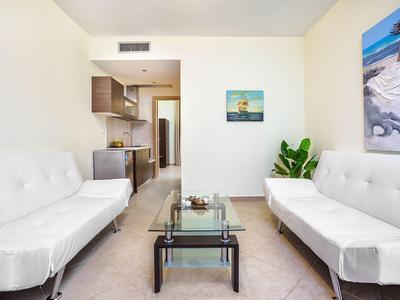 Bright living room with white sofas, glass table, and kitchenette in the background.