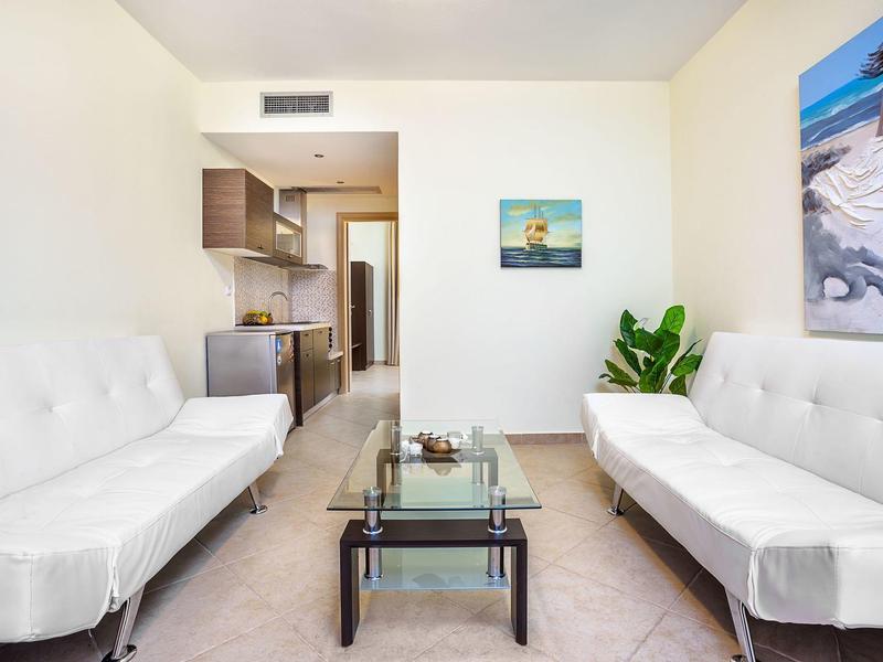 Bright living room with white sofas, glass table, and kitchenette in the background.