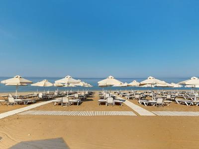 Breiter Sandstrand mit weißen Sonnenschirmen und Liegen, ruhiges Meer, blauer Himmel.