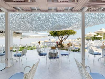 Offene Terrasse eines Hotels mit weißen Tischen und Stühlen unter einem Netzdach mit Blick auf eine Poollandschaft.