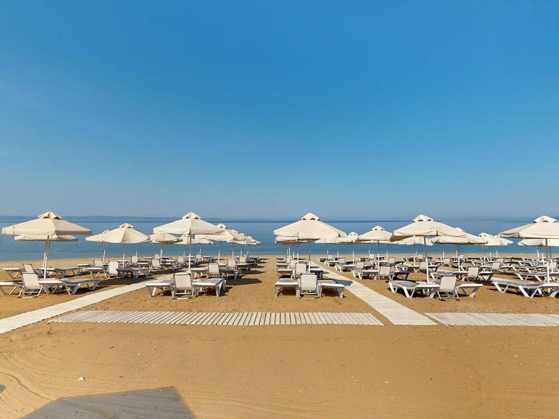 Breiter Sandstrand mit weißen Sonnenschirmen und Liegen, ruhiges Meer, blauer Himmel.