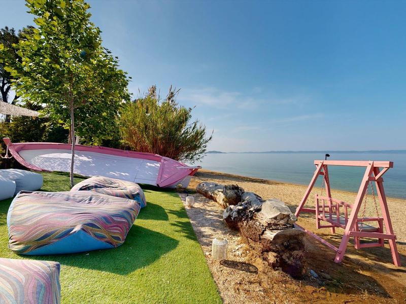 Strand mit grünem Rasen, bunten Sitzkissen, rosa Schaukel und aufgehängten Kajaks am Meer.