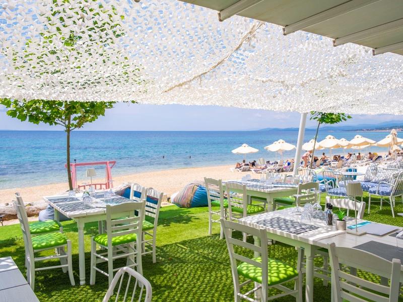 Freier Strandblick mit weißen Stühlen und Tischen unter Sonnensegeln nahe blauem Meer und Sandstrand.