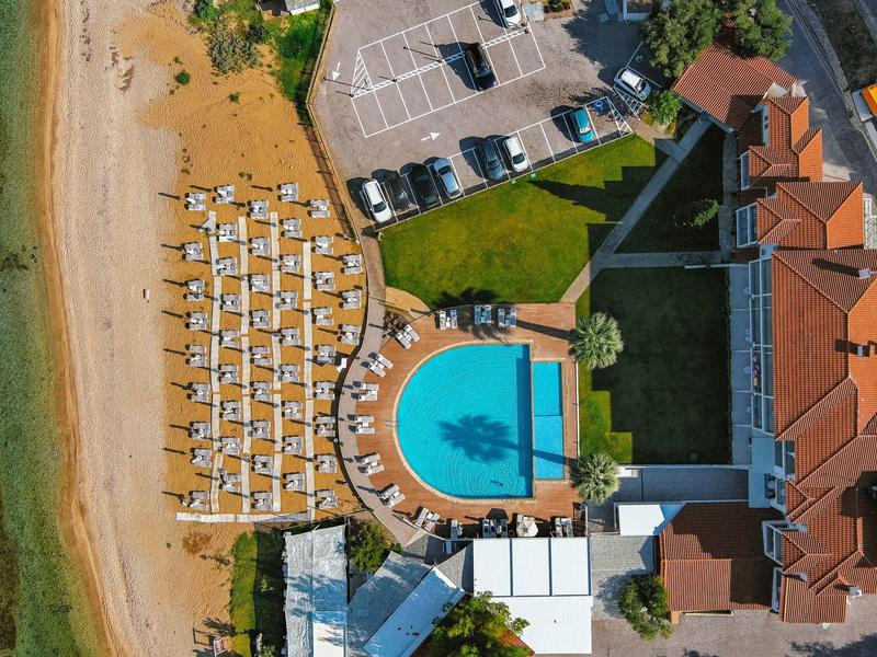Luftaufnahme eines Strandhotels mit Pool, Liegestühlen am Strand und Parkplätzen neben Gebäuden.