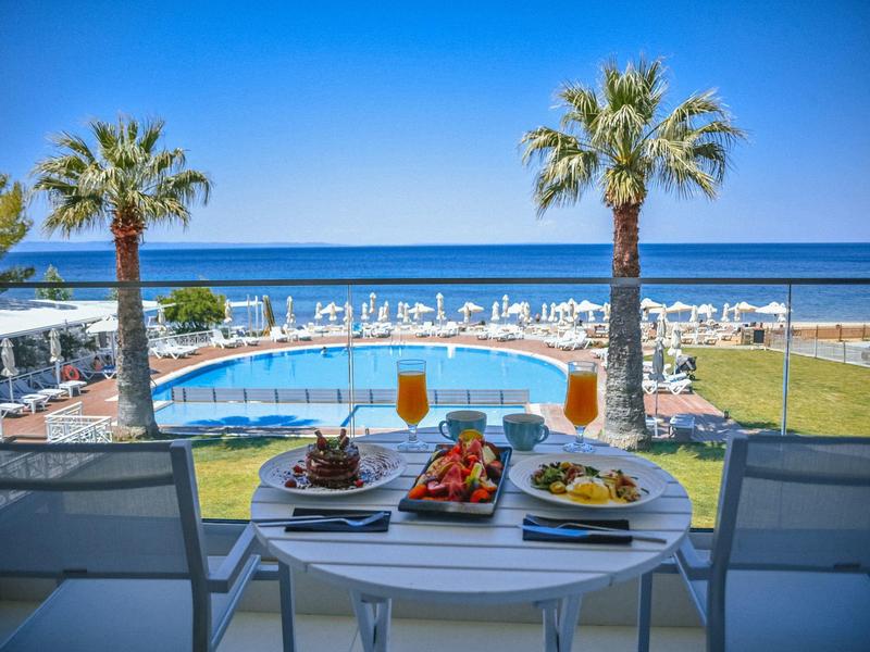 Blick auf Pool und Meer, zwei Drinks und Teller mit Essen auf Tisch, zwei Stühle davor.