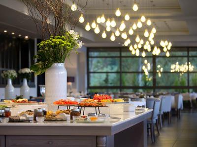 Buffet mit verschiedenen Speisen auf weißem Tisch, moderner Raum mit großen Fenstern und hellem Kronleuchter.