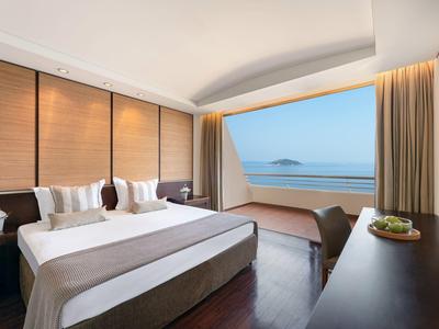 Helles Hotelzimmer mit großem Bett, Holzmöbeln und Panoramafenster mit Meerblick und Insel.