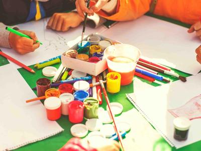Kinderhände malen mit Pinseln und bunten Farben auf Papier am grünen Tisch.