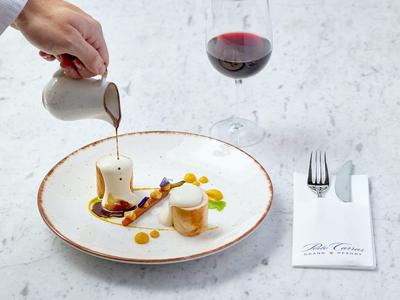 Eine Hand gießt Soße auf ein kunstvoll angerichtetes Dessert auf weißem Teller mit Rotweinglas.