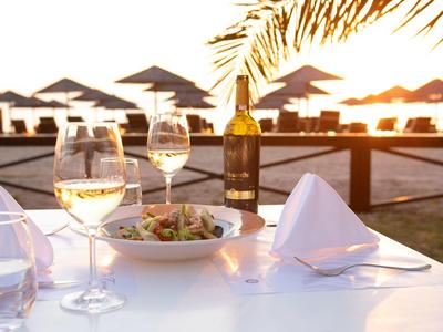 Tisch mit Wein, Salat und Servietten vor Strand mit Sonnenschirmen im Sonnenuntergang.