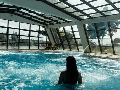 Person im blauen Indoor-Schwimmbad mit Glasdach und großen Fenstern, die Außenbäume zeigen.