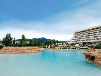 Hotel mit großem Außenpool, Felsen und Bäumen bei klarem Himmel und Bergen im Hintergrund.