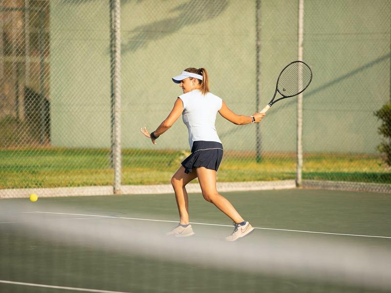 Eine Frau im weißen Shirt spielt Tennis auf einem Freiplatz mit grünem Hintergrund.