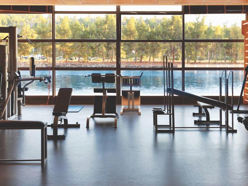 Moderner Fitnessraum mit Geräten, großen Fenstern und Blick auf einen See und Bäume im Hintergrund.