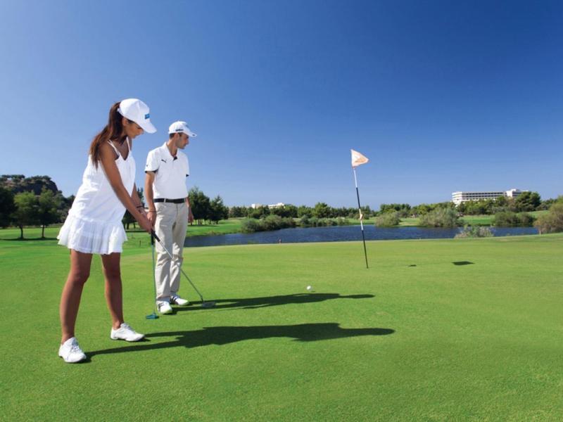 Zwei Golfer auf grünem Platz mit Fahne, klarem Himmel und Wasser im Hintergrund.