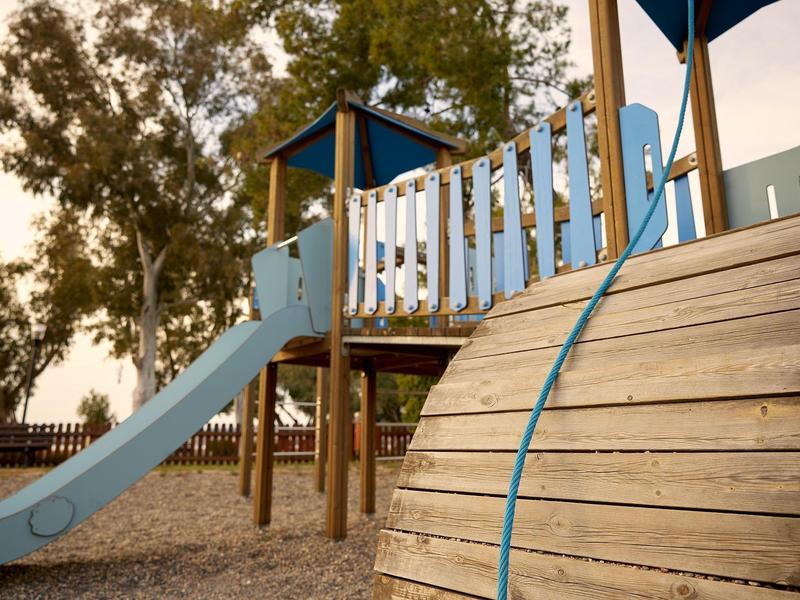 Spielplatz mit blauem Rutsche, Holzklettergerüst und Bäumen im Hintergrund bei Sonnenuntergang.