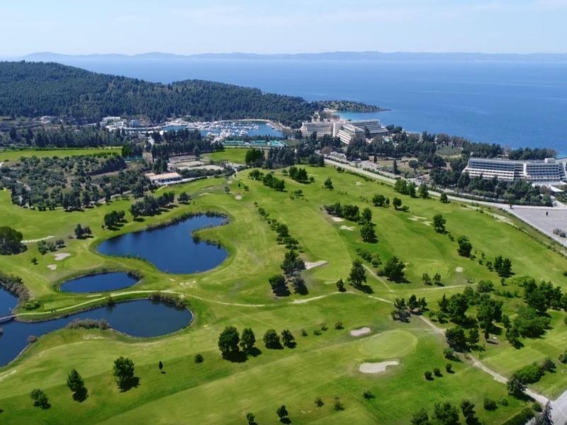 Grüne Golfplätze mit Teichen, Bäumen und Gebäuden nahe Küste und Meer unter blauem Himmel.