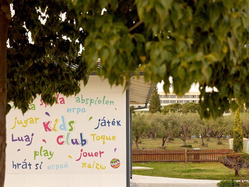 Wand mit bunten Wörtern für ?spielen? in verschiedenen Sprachen, umgeben von grünen Bäumen.