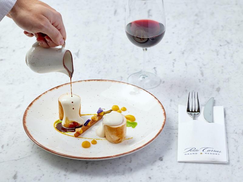 Eine Hand gießt Soße auf ein kunstvoll angerichtetes Dessert auf weißem Teller mit Rotweinglas.