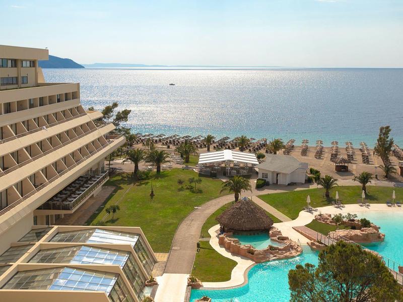 Hotel mit Terrasse, großem Pool, grüner Garten und Blick auf ruhiges Meer bei klarem Himmel.
