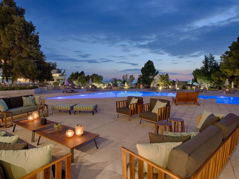 Abendliche Terrasse mit gemütlichen Holzmöbeln und beleuchtetem Pool unter blauem Himmel.