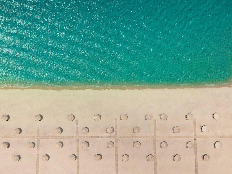 Blick aus der Vogelperspektive auf einen Strand mit Reihen von Sonnenschirmen und türkisfarbenem Meer.