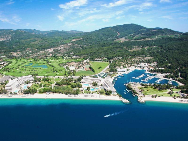 Küstenlandschaft mit blauem Meer, Sandstrand, grünen Feldern und bewaldeten Hügeln im Hintergrund.