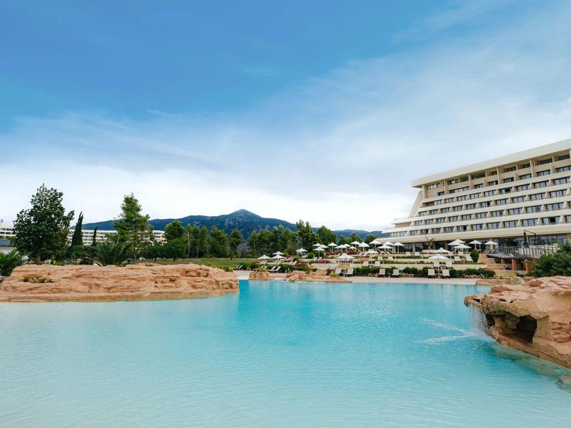 Großer blauer Pool mit Felsenrand, daneben mehrstöckiges Hotel unter blauem Himmel.
