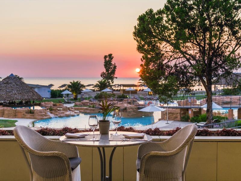 Blick auf Pool, Palmen und Sonnenuntergang von Terrasse mit Tisch, Stühlen und Pflanzen.