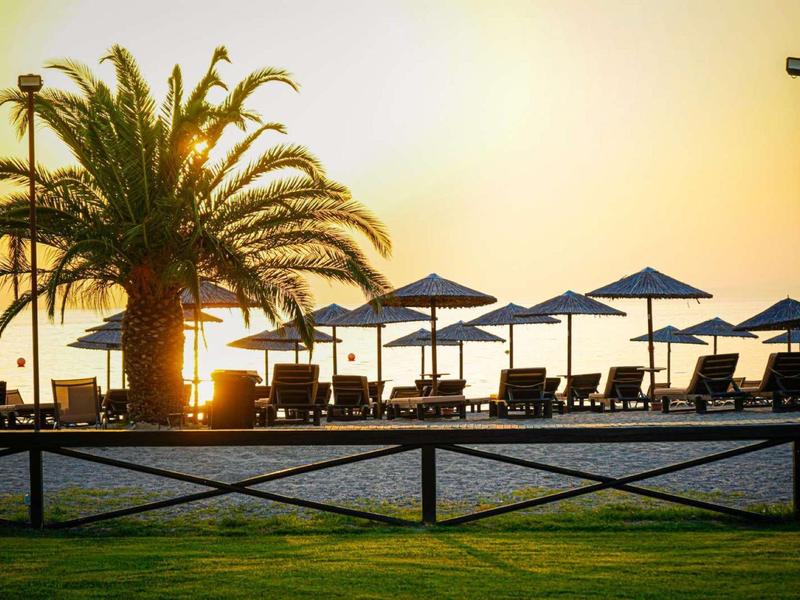 Palmen und Sonnenschirme am Strand bei Sonnenuntergang mit grüner Wiese im Vordergrund.