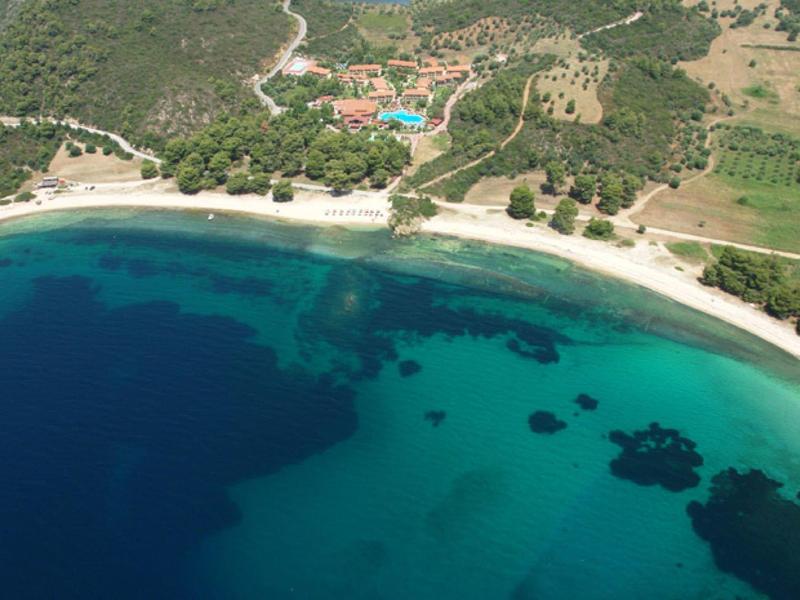 Luftaufnahme einer Bucht mit klarem, blaugrünem Wasser und einem langen Sandstrand, umgeben von grüner Landschaft.